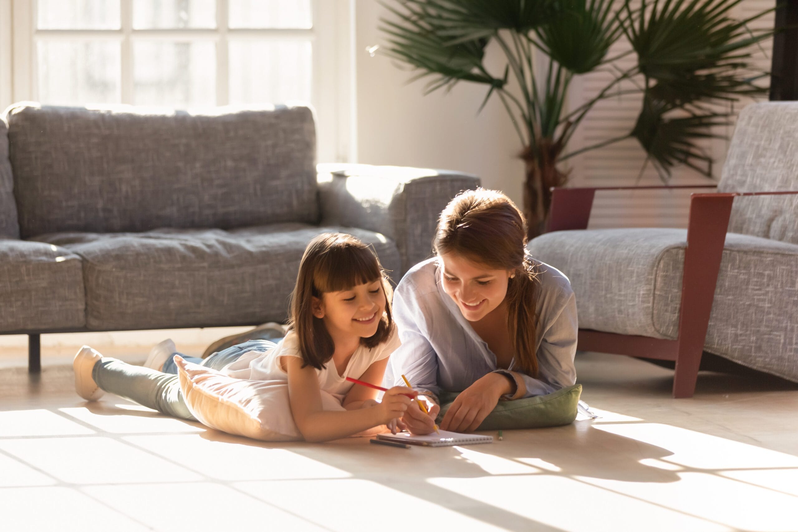 Happy mom helping child daughter drawing with colored pencils laying on warm floor together, smiling baby sitter mother teaching cute kid learning creative activity play laugh at home in living room